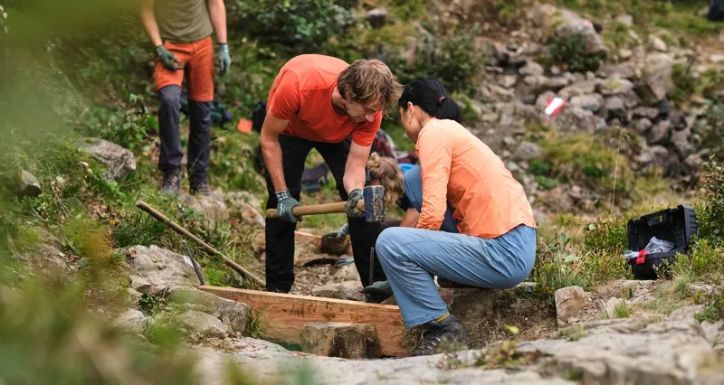 Twee mensen in oranje shirts en handschoenen werken samen op een rotsachtig pad in de buitenlucht; ze gebruiken gereedschap om te graven of te bouwen, terwijl op de achtergrond groen en andere teamleden te zien zijn.